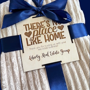 May include: A cream-colored, ribbed blanket tied with a navy blue satin ribbon and bow. A wooden tag with the text "THERE'S NO PLACE LIKE HOME" and "Liberty Real Estate Group" is attached.