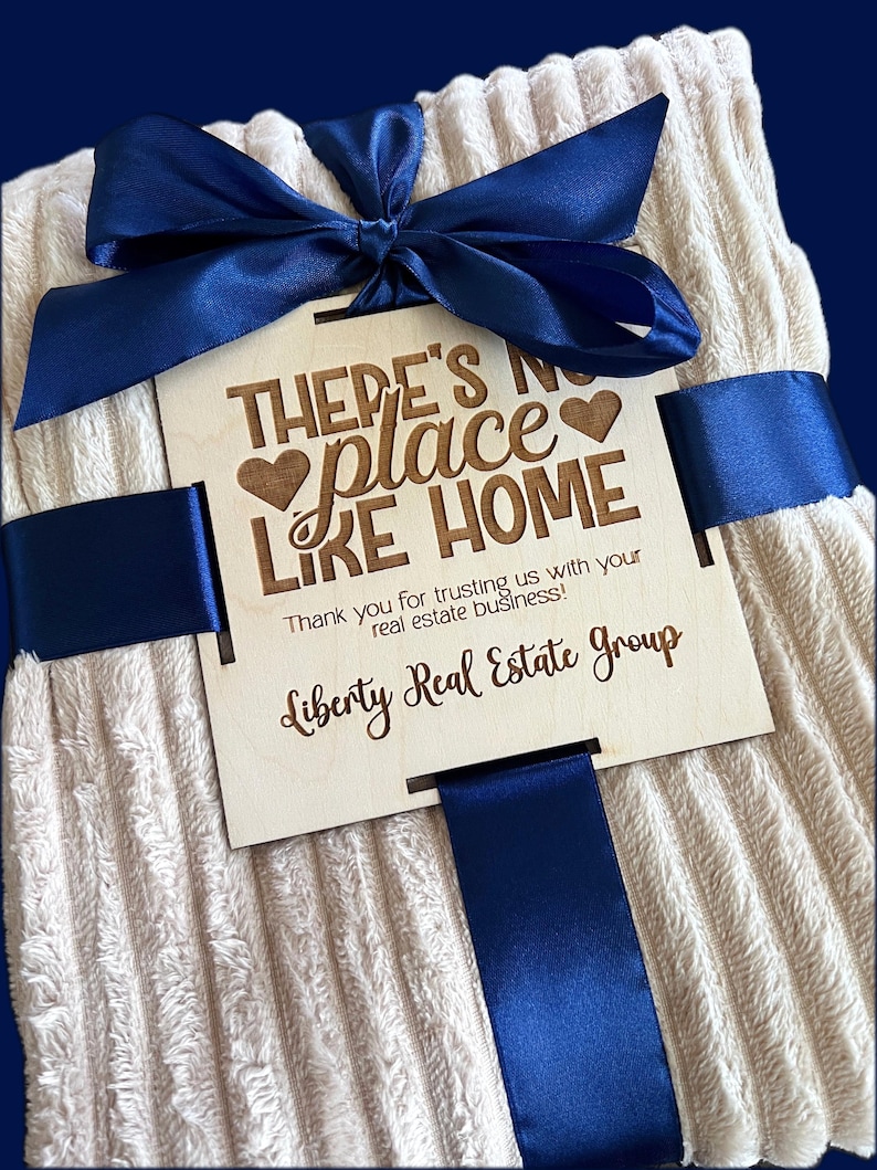 May include: A cream-colored, ribbed blanket tied with a navy blue satin ribbon and bow. A wooden tag with the text "THERE'S NO PLACE LIKE HOME" and "Liberty Real Estate Group" is attached.