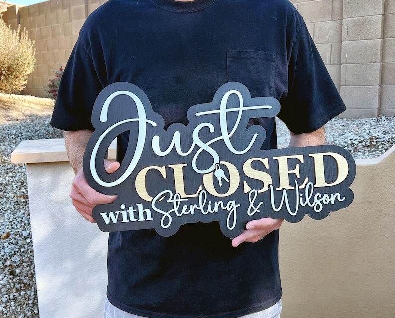 Pu&ograve; includere: Un cartello in legno nero con scritte bianche e dorate che recita Just CLOSED with Sterling & Wilson, con una piccola grafica di due chiavi appese alla lettera O di CLOSED. Il cartello presenta un design a strati con sfondo scuro.