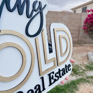 May include: A white wooden sign with the words "Going Gold Real Estate" in black lettering. The letters are layered and have a gold outline.