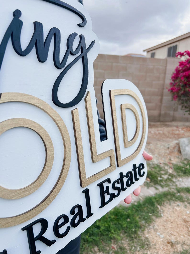 May include: A white wooden sign with the words "Going Gold Real Estate" in black lettering. The letters are layered and have a gold outline.