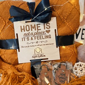 May include: A gift basket with a brown blanket, a wooden sign that says "HOME IS not a place, IT'S A FEELING", and two leather keychains that say "HOME Sweet HOME".