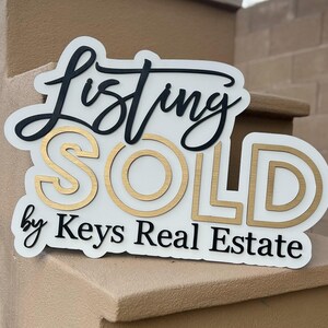 May include: White sign with black and gold lettering that reads "Listing SOLD by Keys Real Estate".