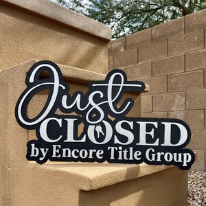 Pu&ograve; includere: Un cartello in bianco e nero con il testo Just CLOSED by Encore Title Group in uno stile a caratteri sovrapposti in corsivo e stampatello. La parola CLOSED include una piccola grafica di una chiave di casa integrata nella lettera O.