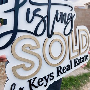 May include: A white wooden sign with black and gold lettering that reads "Listing Sold by Keys Real Estate".