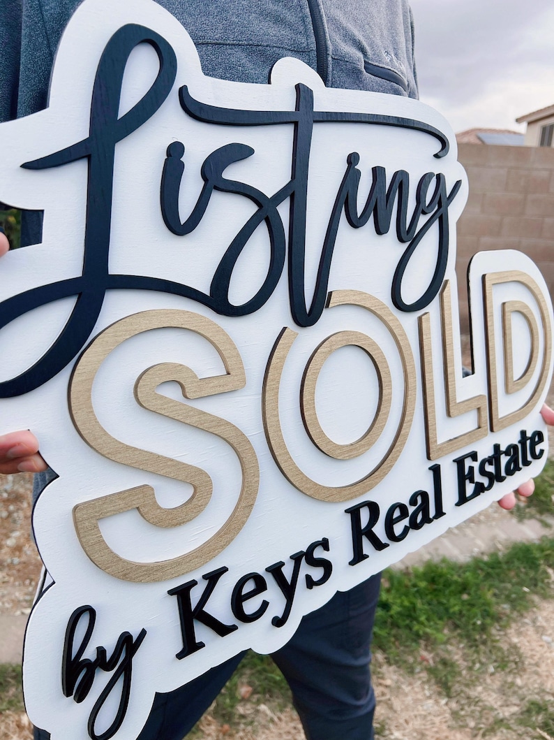May include: A white wooden sign with black and gold lettering that reads "Listing Sold by Keys Real Estate".