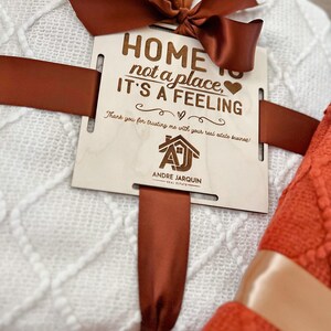 May include: A white and orange blanket set with a wooden sign that reads "Home is not a place, it's a feeling." The sign also includes the text "Thank you for trusting me with your real estate business!" and a logo for Andre Jarquin Real Estate.