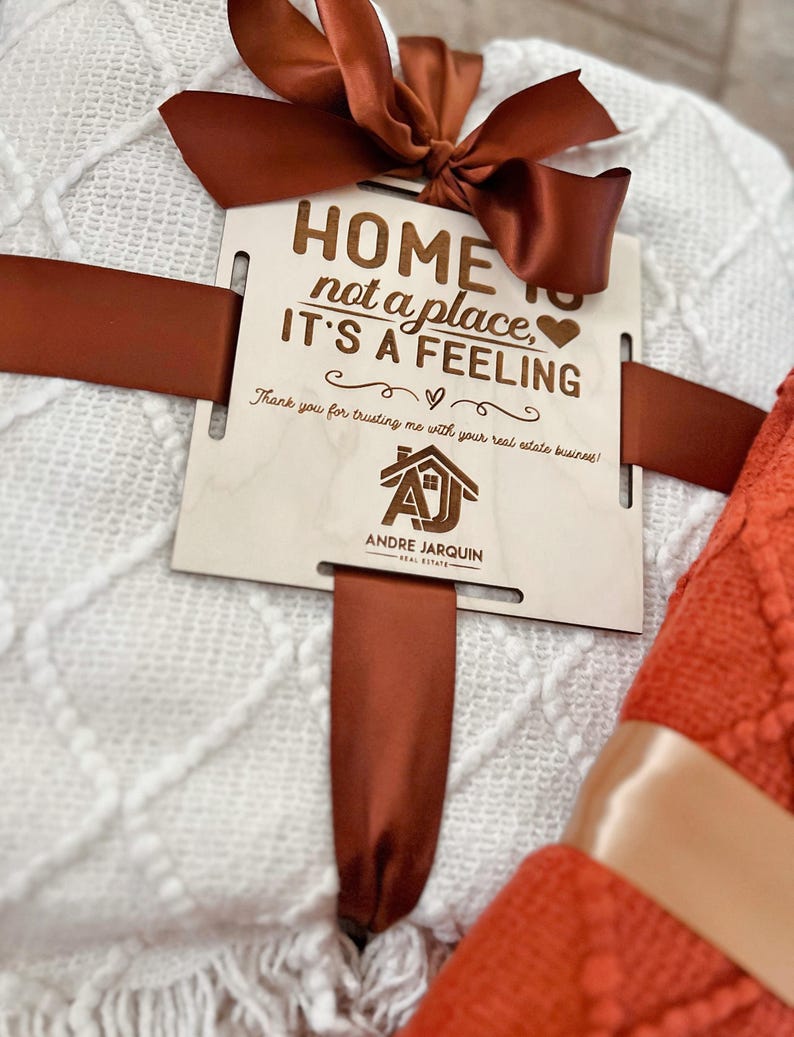 May include: A white and orange blanket set with a wooden sign that reads "Home is not a place, it's a feeling." The sign also includes the text "Thank you for trusting me with your real estate business!" and a logo for Andre Jarquin Real Estate.