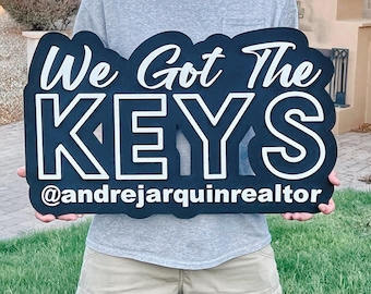 May include: A black and white wooden sign with the text "We Got The KEYS" and the Instagram handle "@andrejarquinrealtor". The sign is held by a person in a gray shirt.