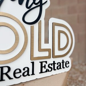 May include: A white sign with the words "Going Gold Real Estate" in black and gold lettering. The letters are raised and have a textured finish.