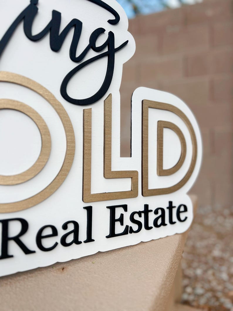 May include: A white sign with the words "Going Gold Real Estate" in black and gold lettering. The letters are raised and have a textured finish.