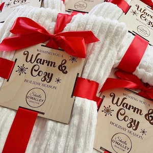 May include: White, textured towels tied with red satin ribbon and wooden tags. The tags read "Wishing You a Warm & Cozy Holiday Season" and include a realtor's name. A festive gift set.