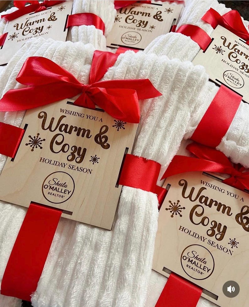 May include: White, textured towels tied with red satin ribbon and wooden tags. The tags read "Wishing You a Warm & Cozy Holiday Season" and include a realtor's name. A festive gift set.