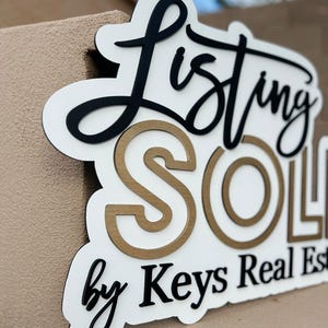 May include: White sign with black lettering that reads "Listing Sold by Keys Real Estate". The word "Sold" is in gold lettering with a white outline.