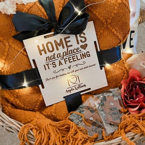 May include: A wicker basket filled with a cozy orange throw blanket, a wooden sign that reads "HOME IS not a place, IT'S A FEELING", and a small house-shaped keychain with the text "HOME SWEET HOMES Angela Williams".