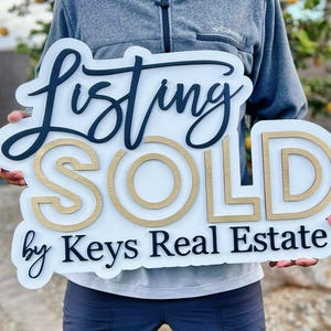 May include: White wooden sign with black lettering that reads "Listing SOLD by Keys Real Estate". The word "SOLD" is in gold lettering with a double outline.