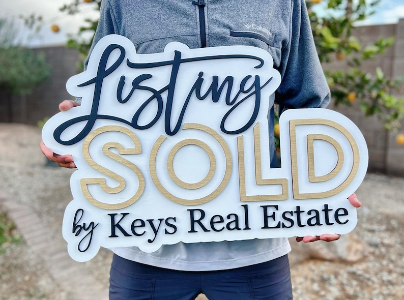 May include: White wooden sign with black lettering that reads "Listing SOLD by Keys Real Estate". The word "SOLD" is in gold lettering with a double outline.