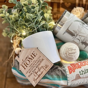 May include: A wooden house-shaped welcome sign with the text "Welcome Home" and a message thanking a realtor for their business. The sign is sitting on a woven basket with a small potted plant, a white tumbler, and a decorative ball.
