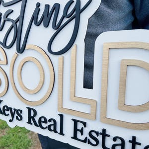 May include: A white wooden sign with the words "Just Sold" in black and gold lettering. The sign also includes the text "Keys Real Estate" in black lettering.