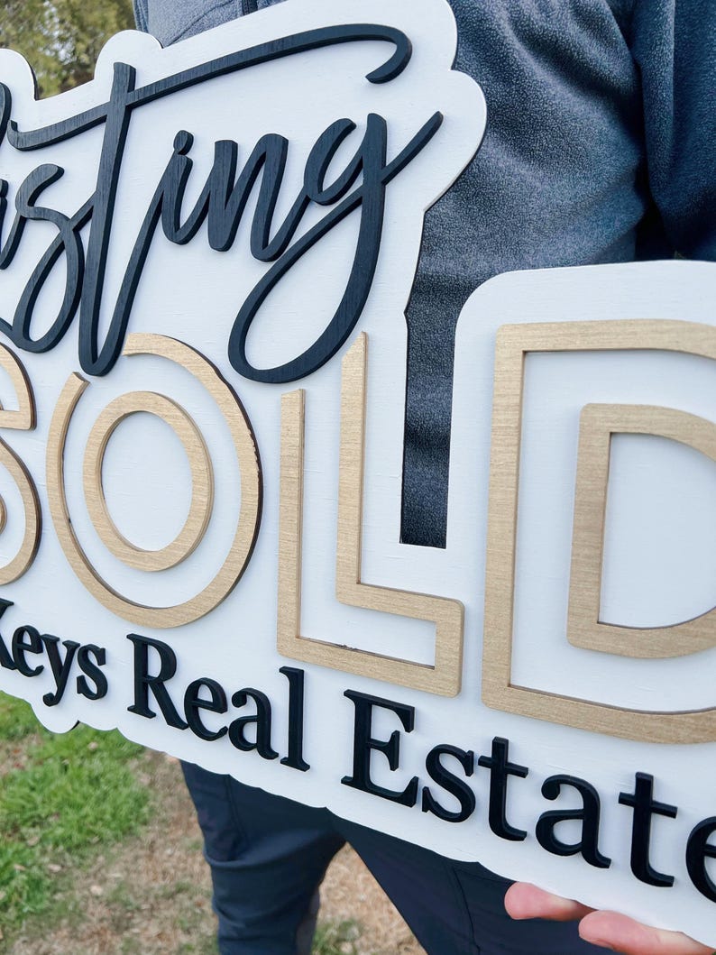 May include: A white wooden sign with the words "Just Sold" in black and gold lettering. The sign also includes the text "Keys Real Estate" in black lettering.