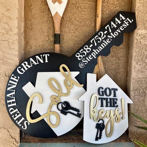 May include: A large black and white wooden key-shaped sign featuring the text Stephanie Grant, 858-752-7444, and @StephanieMovesFL. A smaller white house-shaped sign displays the text Sold and Got the keys! with black key icons.