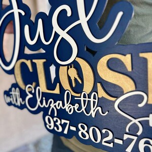 Pu&ograve; includere: Un'insegna in legno a strati con il testo Just Closed with Elizabeth S e il numero di telefono 937-802-76. L'insegna presenta una base blu scuro, scritte bianche in corsivo e lettere dorate con un'icona a forma di chiave nella lettera O.
