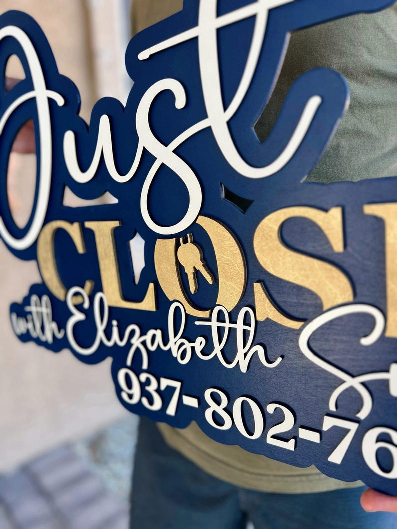 Pu&ograve; includere: Un'insegna in legno a strati con il testo Just Closed with Elizabeth S e il numero di telefono 937-802-76. L'insegna presenta una base blu scuro, scritte bianche in corsivo e lettere dorate con un'icona a forma di chiave nella lettera O.