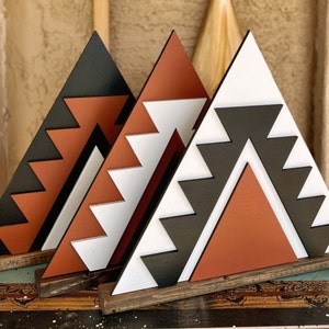 May include: Three wooden triangle wall decor pieces with geometric patterns in black, white, and orange. The triangles are stacked on top of each other and have a rustic wood base.