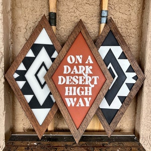 May include: Three diamond-shaped wooden signs with black and white geometric patterns. The center sign has a red background with the text "ON A DARK DESERT HIGHWAY" in white.