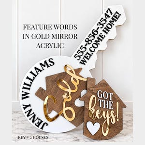 May include: A large white key-shaped sign with black text reading 555-856-3547 WELCOME HOME, paired with two house-shaped wooden signs. One house features the name JENNY WILLIAMS and gold mirror acrylic text, while the other says GOT THE keys!.
