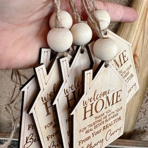 May include: Four wooden house-shaped welcome home signs with twine and wooden beads. The signs have a welcome home message and the text "Thank you for trusting me with your real estate business! From your realtor, Bethany Curry".