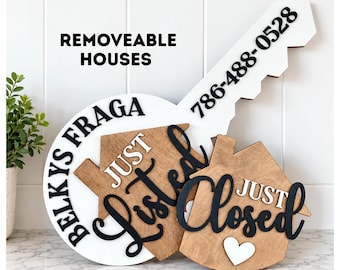 May include: A white key-shaped sign with black text reading REMOVEABLE HOUSES, BELKYS FRAGA, and 786-488-0528. Two wooden house-shaped plaques are attached, one with black text JUST LISTED and the other with black text JUST CLOSED and a white heart.