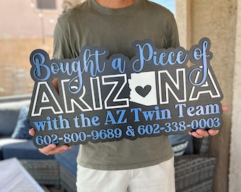 May include: A wooden sign with the words "Bought a Piece of ARIZONA" in blue and white lettering. The sign features the Arizona state outline with a heart, and the text "with the AZ Twin Team" and phone numbers.