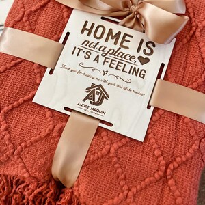 May include: A red throw blanket with a fringe border is tied with a brown ribbon and a wooden sign that reads "Home is not a place, it's a feeling." The sign also includes the text "Thank you for trusting me with your real estate business!" and a logo for Andre Jarquin Real Estate.