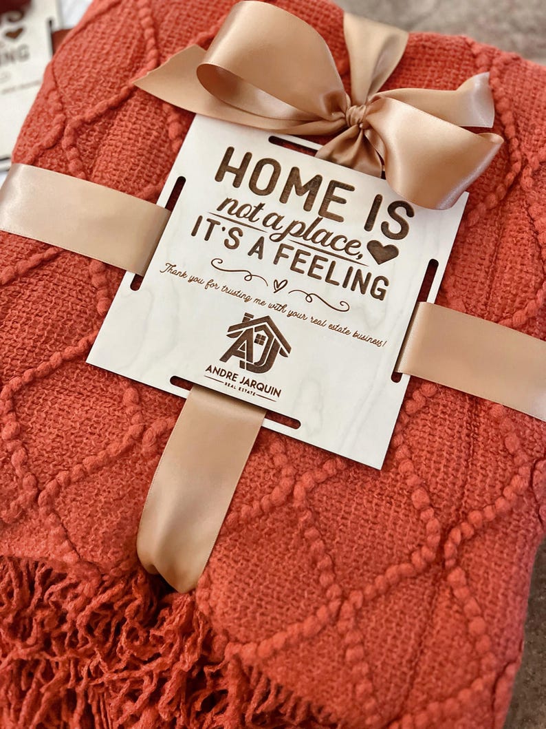 May include: A red throw blanket with a fringe border is tied with a brown ribbon and a wooden sign that reads "Home is not a place, it's a feeling." The sign also includes the text "Thank you for trusting me with your real estate business!" and a logo for Andre Jarquin Real Estate.