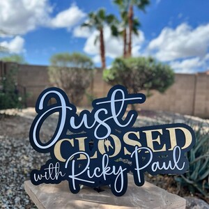 Pu&ograve; includere: Un cartello nero e oro con scritte bianche e dorate che recita Just CLOSED with Ricky Paul. Il cartello presenta una sagoma di una chiave di casa integrata nella parola CLOSED ed &egrave; progettato con una combinazione di caratteri corsivi e stampatello.