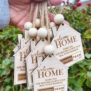 May include: A set of wooden house-shaped welcome home tags with the text "Welcome Home" and "Thank you for trusting us with your real estate business!"  The tags are tied together with twine and wooden beads. The text "Bowers Team" is also included on the tags.