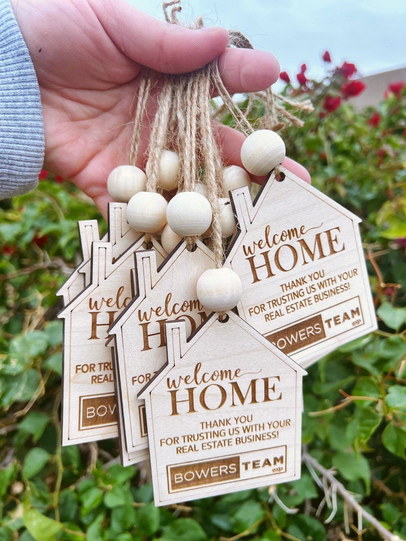 May include: A set of wooden house-shaped welcome home tags with the text "Welcome Home" and "Thank you for trusting us with your real estate business!"  The tags are tied together with twine and wooden beads. The text "Bowers Team" is also included on the tags.