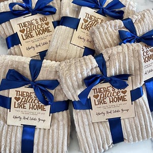 May include: Several beige, corduroy-textured blankets are bundled with navy blue satin ribbon and bows. Each blanket has a wooden tag with the text "THERE'S NO PLACE LIKE HOME" and "Liberty Real Estate Group."