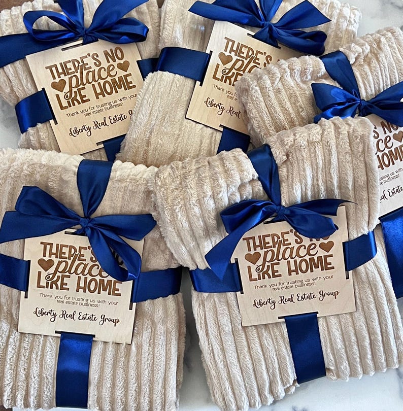 May include: Several beige, corduroy-textured blankets are bundled with navy blue satin ribbon and bows. Each blanket has a wooden tag with the text "THERE'S NO PLACE LIKE HOME" and "Liberty Real Estate Group."