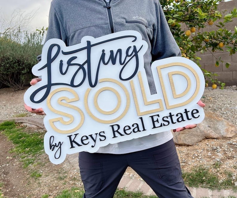 May include: A white sign with black and gold lettering that reads "Listing SOLD by Keys Real Estate".