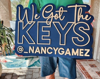 May include: A large, dark blue wooden sign with light-coloured, raised lettering that reads "We Got The KEYS @_NANCYGAMEZ". The sign features a script font for the top line and a bold, block-style font for the middle and bottom lines.