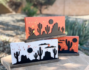 May include: Three rectangular wooden signs featuring desert landscapes. Two signs have orange backgrounds with paint splatters, and one has a white background with orange splatters. Each features black silhouettes of cacti and rock formations.