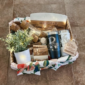 May include: A wicker basket filled with housewarming gifts, including a wooden chopping board with the text "Richard & Kay Home Sweet Home Est. 2023", two personalised tumblers, a small potted plant, and a welcome home sign.