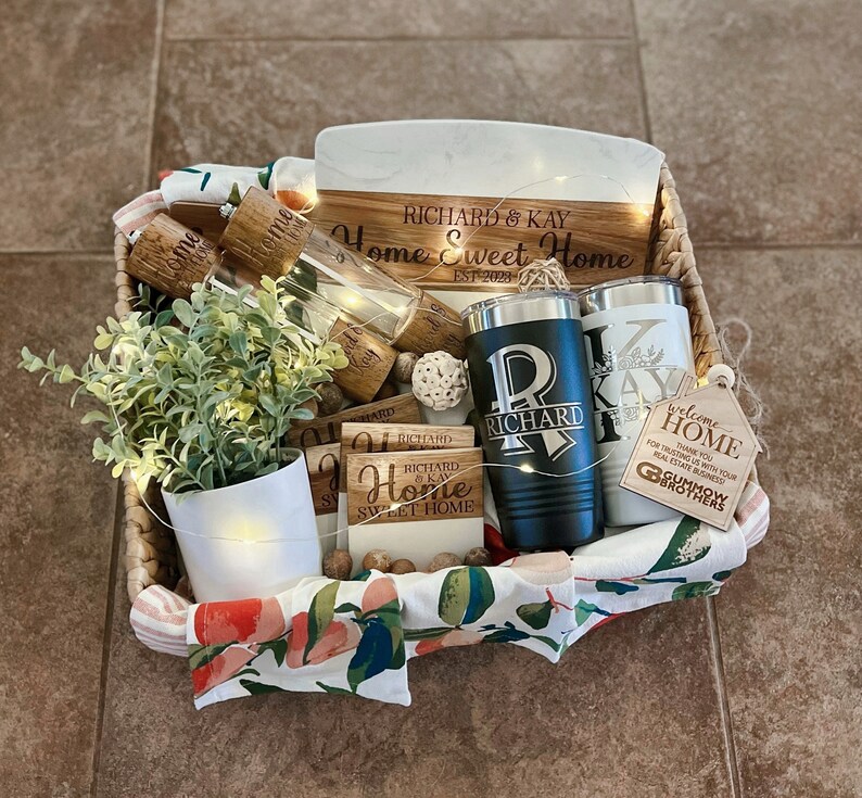 May include: A wicker basket filled with housewarming gifts, including a wooden chopping board with the text "Richard & Kay Home Sweet Home Est. 2023", two personalised tumblers, a small potted plant, and a welcome home sign.