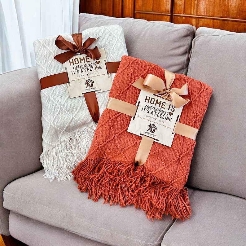 May include: Two knitted throw blankets with fringe, one white and one orange, are tied with ribbon and feature a wooden sign with the text "Home is not a place, it's a feeling".