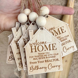 May include: Set of four wooden house-shaped welcome home tags with the text "Welcome Home" and "Thank you for trusting me with your real estate business! From your realtor, Bethany Curry".