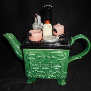 May include: Green ceramic teapot shaped like a vintage stove with a black top and a variety of miniature kitchen accessories, including a kettle, a pot, and a frying pan.