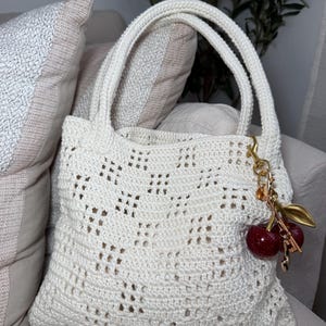 May include: Cream-colored crochet tote bag with a decorative openwork pattern. The bag has two handles and a gold-colored charm with red cherry accents. The bag is sitting on a beige sofa with decorative pillows.