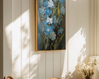Puede incluir: Un cuadro rectangular vertical en un marco dorado fino que representa un grupo de flores nomeolvides de color azul claro con tallos y hojas verdes sobre un fondo oscuro y apagado, expuesto en una pared de paneles blancos sobre una mesa redonda.
